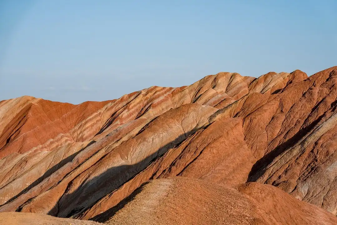 Crestas de montaña de roca con franjas rojizas, ocres y blanquecinas, iluminadas por el sol bajo un cielo azul despejado
