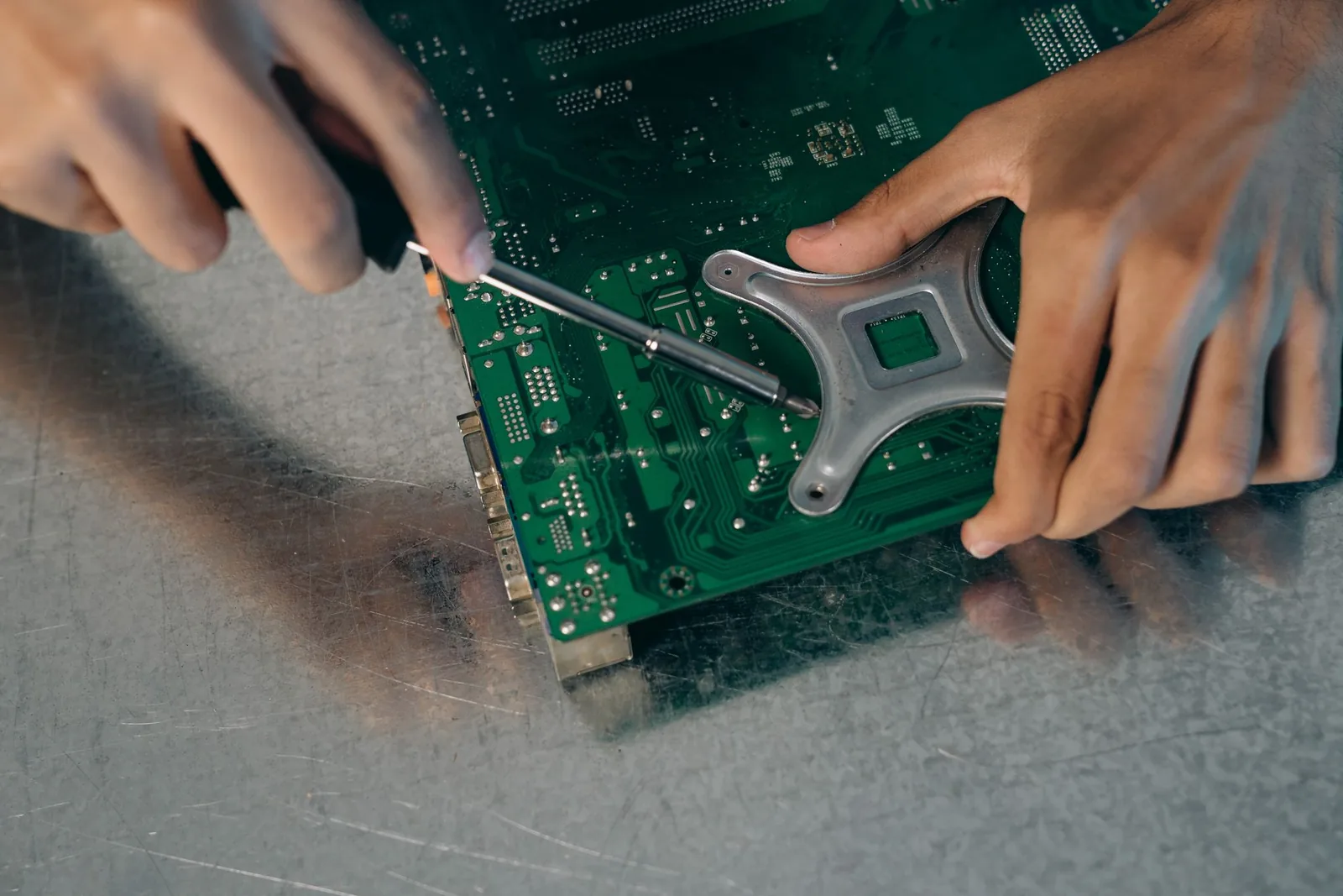 Dos manos con un destornillador sobre una placa de circuito verde apoyada en una mesa de metal rayada