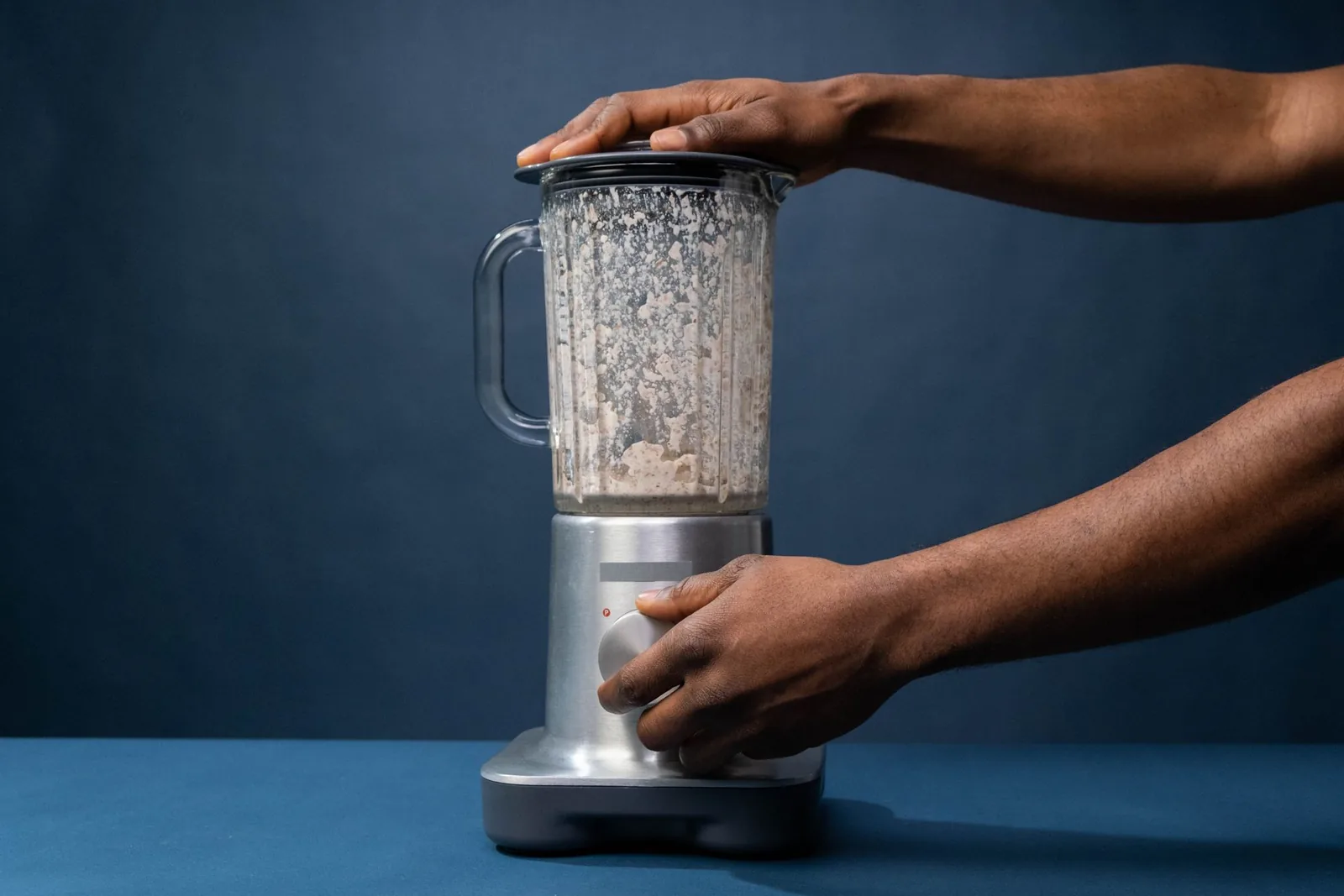 Manos sobre una batidora de vaso metálica con restos de masa en el cristal, un electrodoméstico de cocina de uso diario