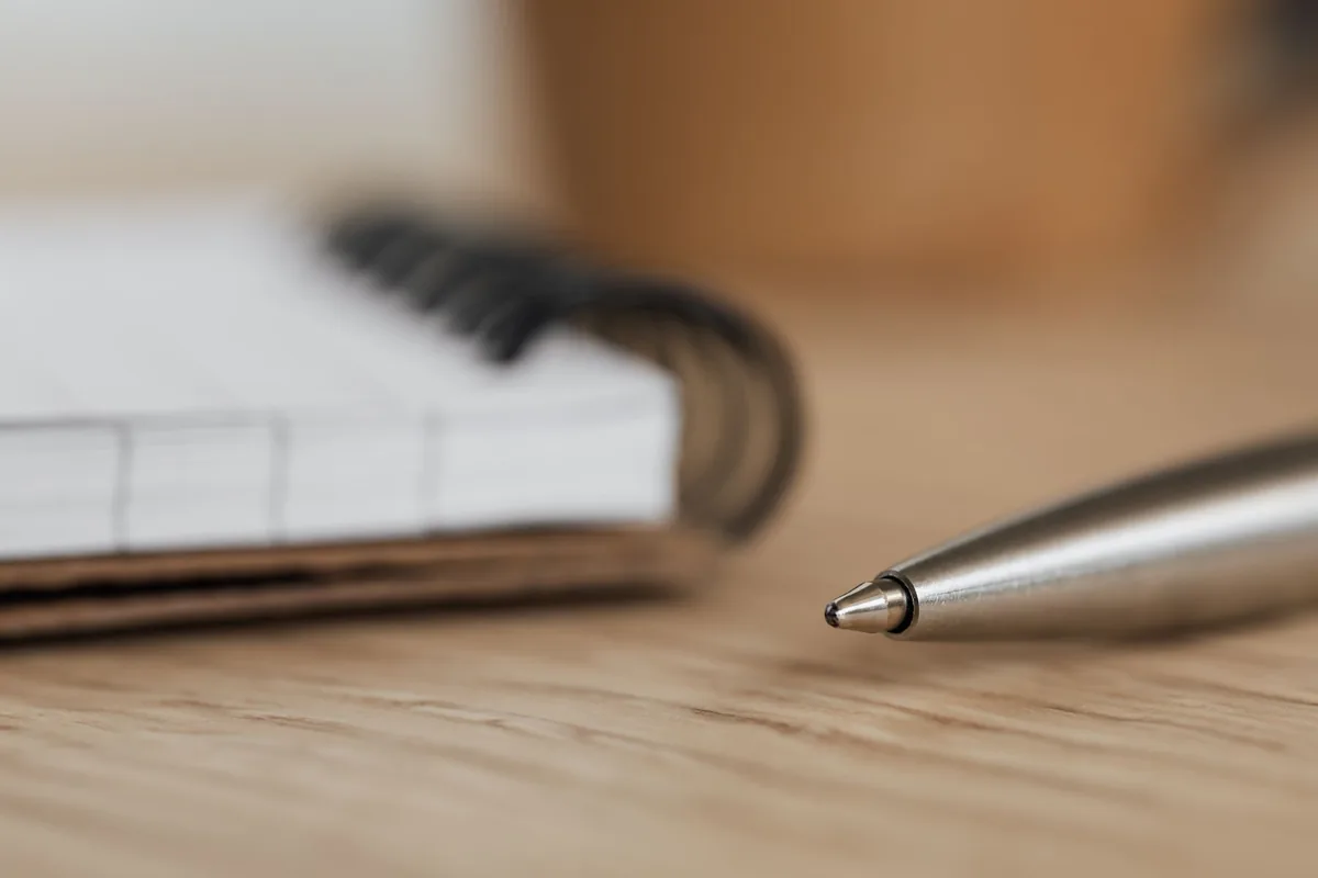 Punta de un bolígrafo metálico junto a un cuaderno de espiral sobre una mesa de madera, en primer plano