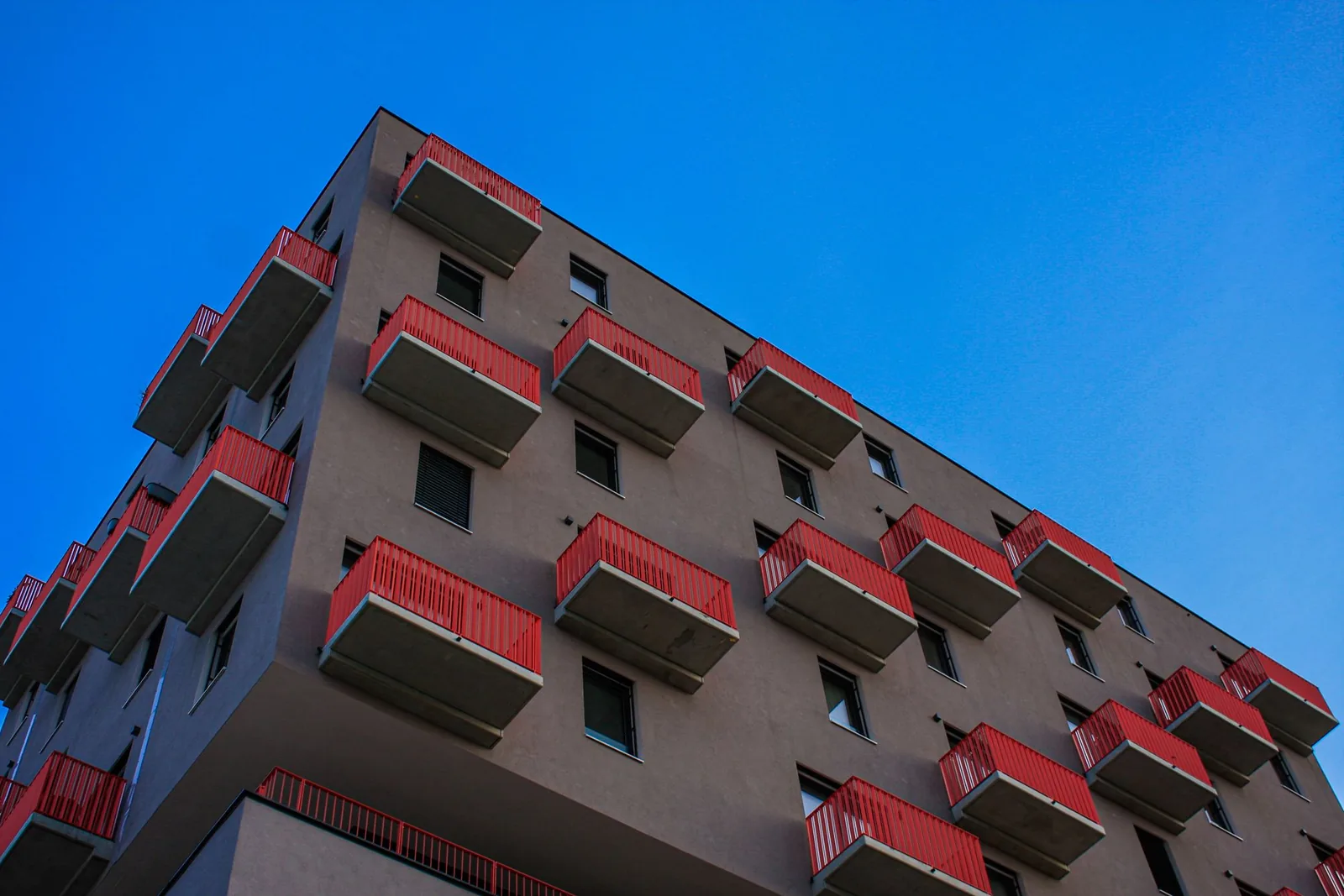 Edificio de pisos de varias plantas visto desde abajo, con balcones de barandilla roja contra un cielo azul