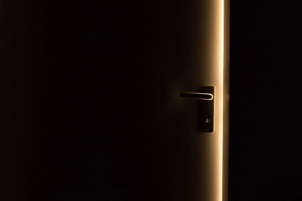 Puerta de una vivienda entreabierta en la penumbra, con una línea de luz y la manija iluminada