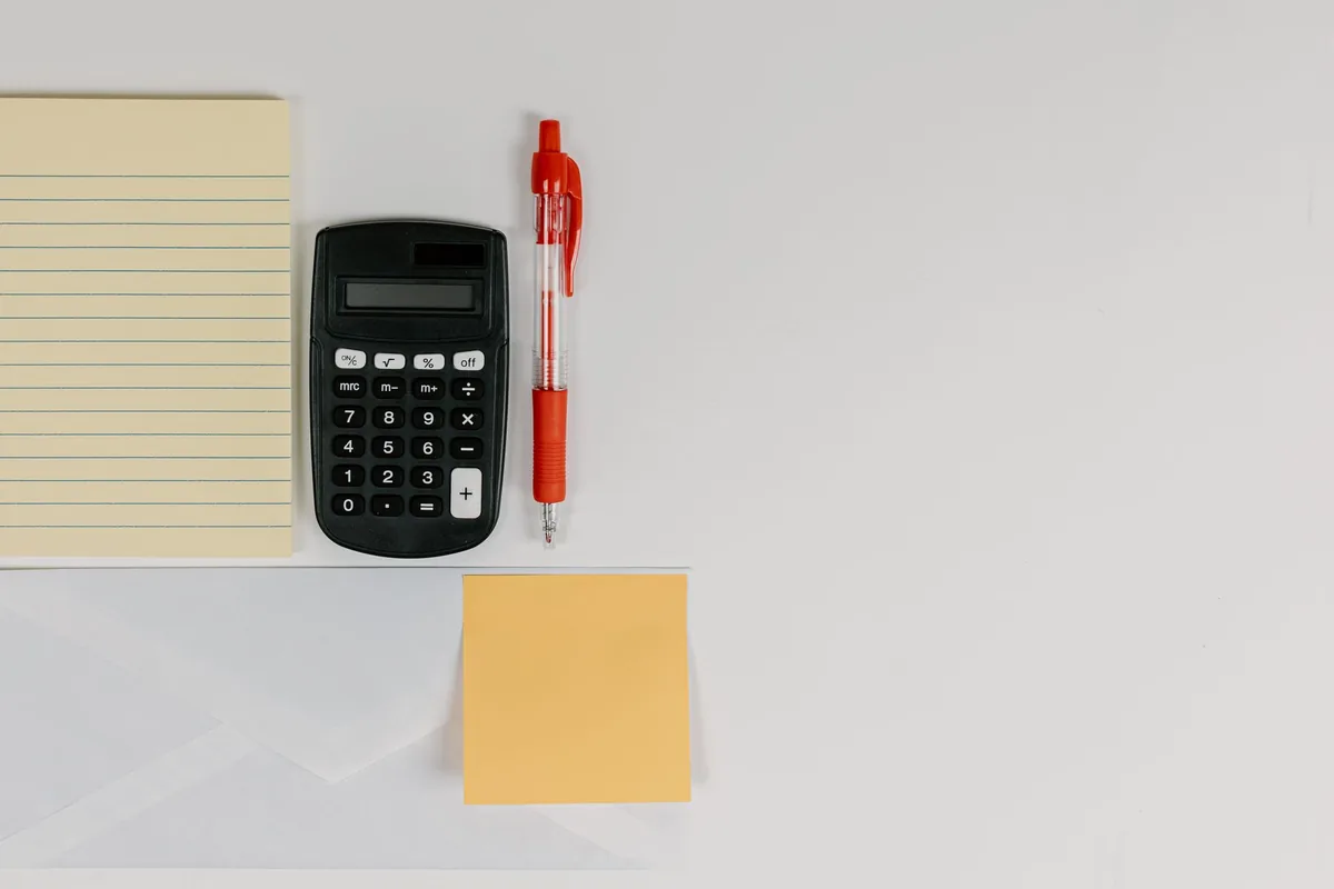 Calculadora negra junto a un bolígrafo rojo, un bloc de notas y una nota adhesiva sobre una mesa blanca