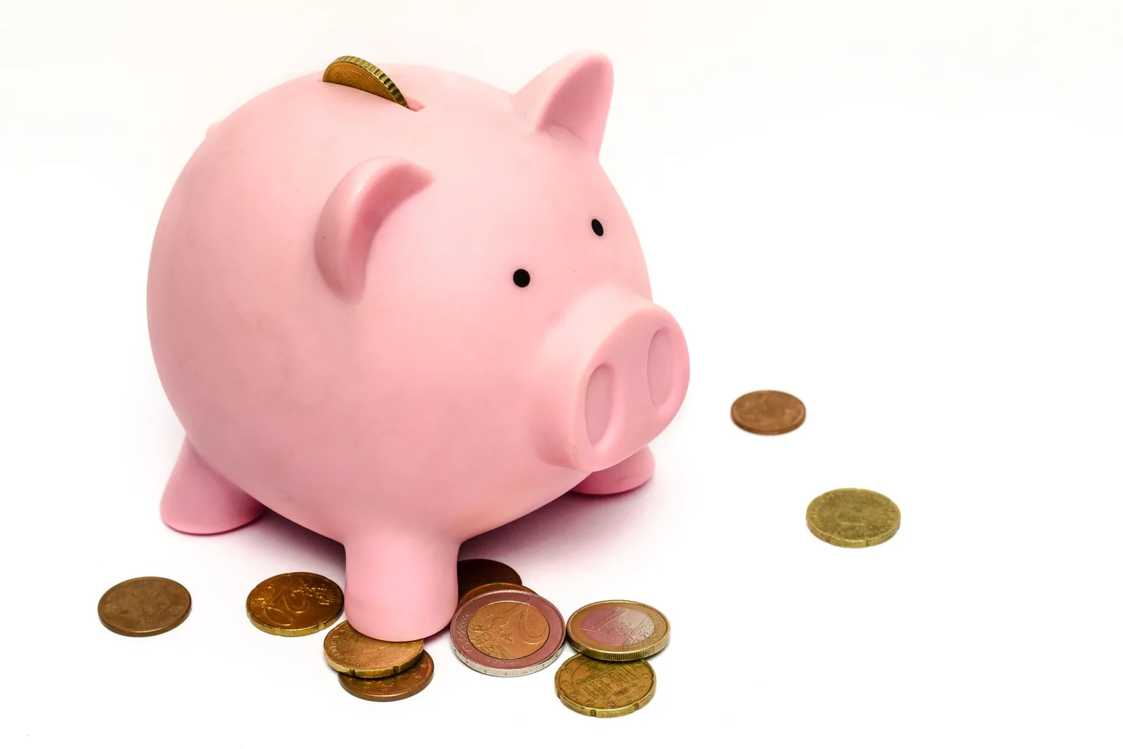 Hucha rosa de cerdito sobre fondo blanco, con una moneda en la ranura y monedas de euro esparcidas alrededor